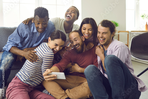 Cheerful company of multiracial friends have fun and take selfies during friendly gatherings at home. People sitting in the living room on the floor capture their moments in life on a mobile phone. © Studio Romantic