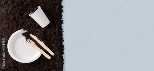 Paper plate and glass  wooden knife and fork on soil on blue background flat lay composition  top view. Compostable or biodegradable dinnerware concept horizontal banner format with copy space