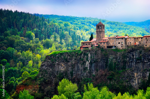 Castellfollit de la Roca, Garrotxa, Province of Girona, Catalonia, Spain, Europe. Beautiful view, ancient church and town houses at the top of huge basalt cliff and mountain covered with green wood photo