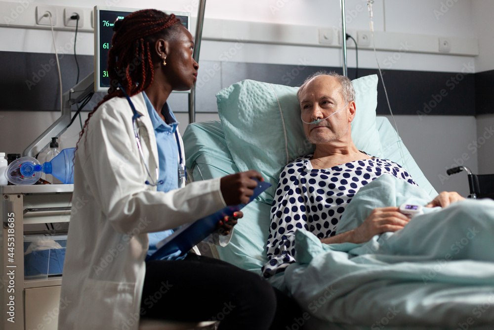 African american doctor reading diagnosis from clipboard to sick ill unwell senior patient laying in bed, breathing with help from oxygen mask, listening discussing with medical staff about recovery. African american doctor reading diagnosis from clipboard to sick ill unwell senior patient laying in bed, breathing with help from oxygen mask, listening discussing with medical staff about recovery.