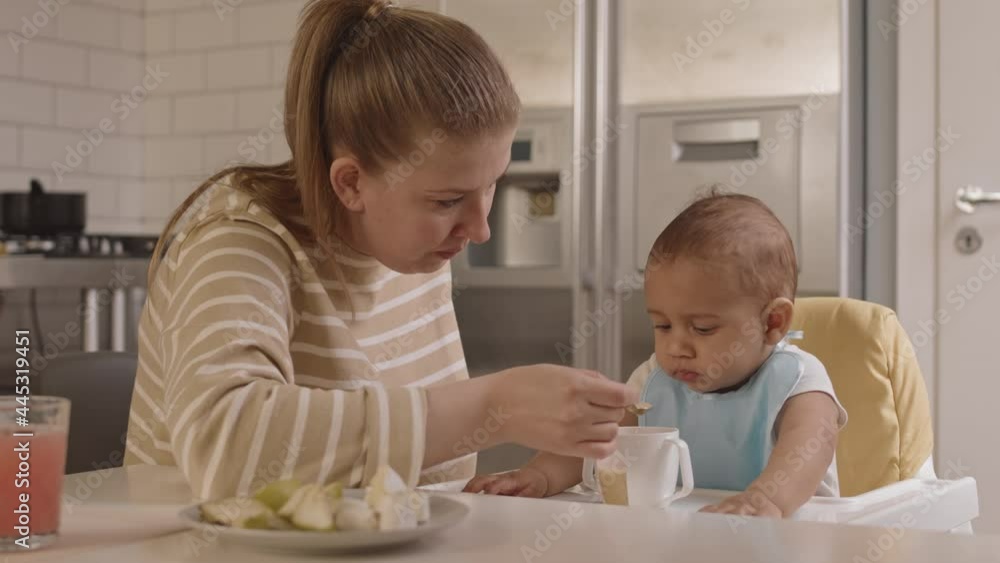 Medium close-up of young Caucasian woman sitting at table, feeding her adorable Mixed-Race child with baby food in kitchen Medium close-up of young Caucasian woman sitting at table, feeding her adorable Mixed-Race child with baby food in kitchen