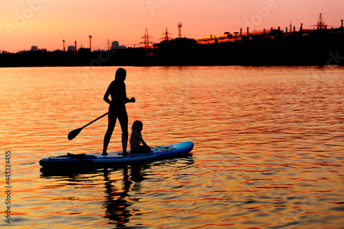 Mom and daughter are surfing on a paddle board at sunset