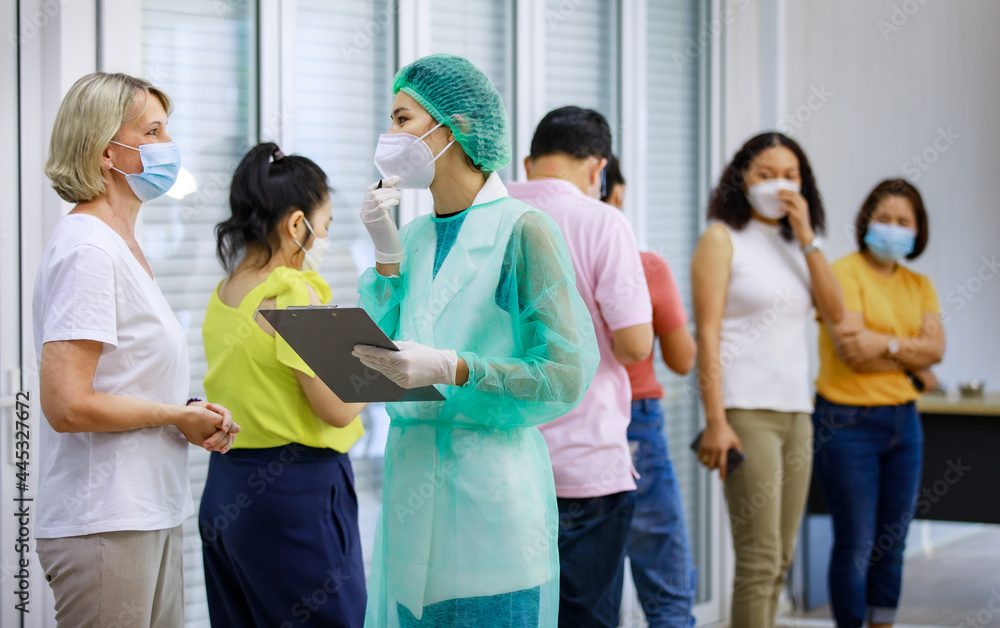 Young female nurse in full hazard protection uniform holding paper board asking personal information from senior caucasian woman patient while others wait in queue line in blurred background Young female nurse in full hazard protection uniform holding paper board asking personal information from senior caucasian woman patient while others wait in queue line in blurred background