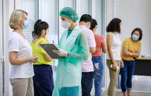 Young female nurse in full hazard protection uniform holding paper board asking personal information from senior caucasian woman patient while others wait in queue line in blurred background