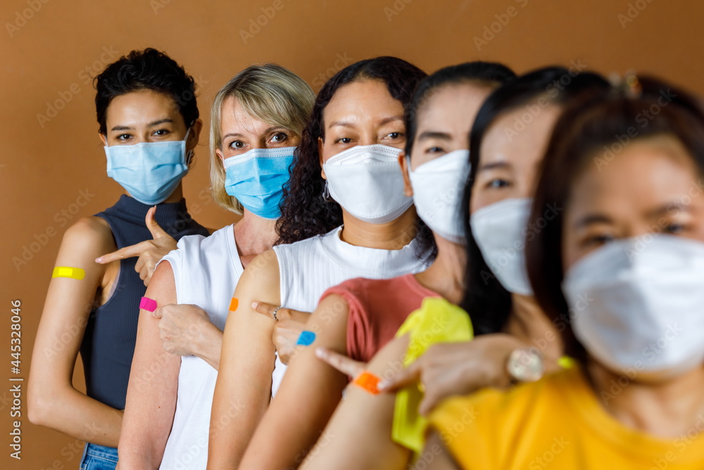 Diverse group of multinational ethnic female patients wears face mask stand in line by height order look at camera pointing colorful plasters on shoulder together after vaccinated