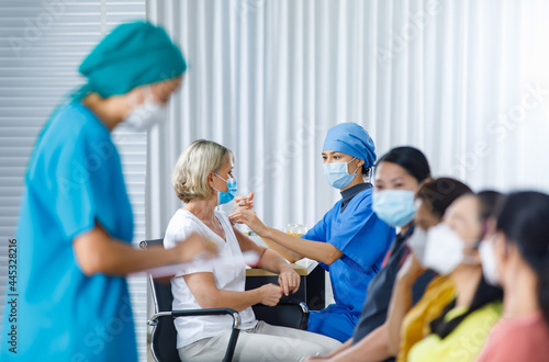 Female doctor wears face mask and hospital uniform injecting coronavirus vaccine into Caucasian senior woman shoulder while other multinational patients wait in queue in blurred foreground