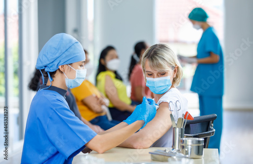 Asian female doctor wears face mask and blue hospital uniform injecting vaccine to Caucasian senior woman at ward working desk with equipment while other patients wait in queue in blurred background