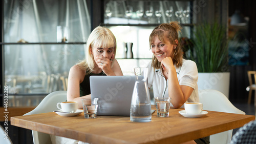 Two Female Colleagues Working Together Feeling Surprised and Humoured by Information on Laptop in a Modern Restaurant Behind a Wooden Desk With Glasses of Water and Coffee Cups. photo