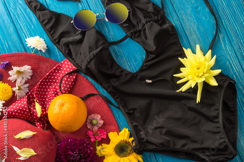 Summer still life with flowers, hat, orange, sunglasses and black swimsuit on blue boards. Background