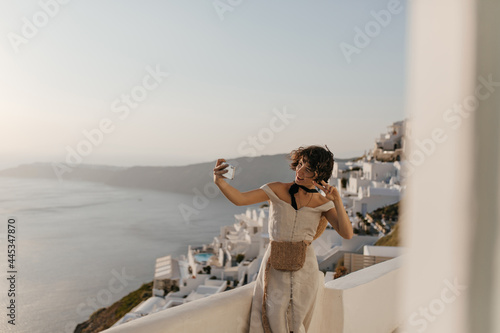 Brunette charming woman in beige stylish summer dress with straw little bag takes selfie on sea and city background and shows v-sign.