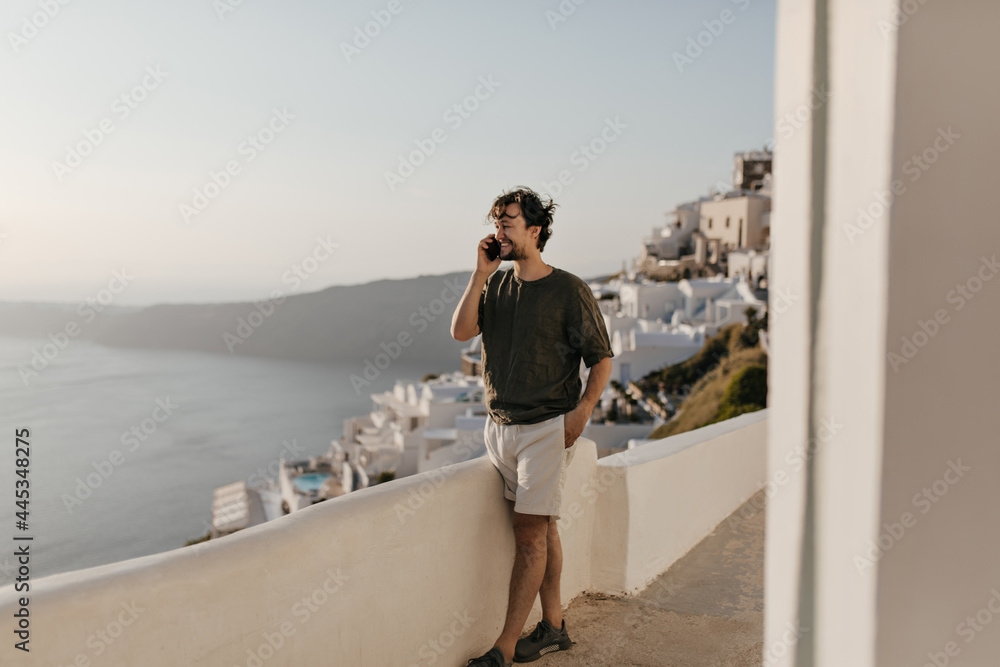 Charming man in white shorts, dark green t-shirt smiles and talks on phone outside. Good humored guy enjoys sea view.