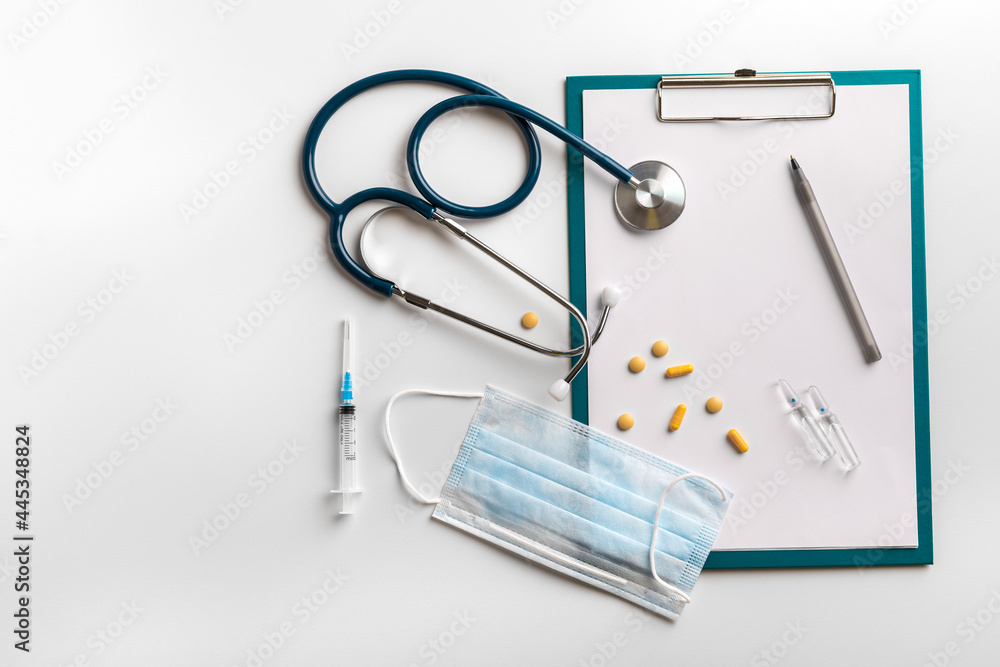 The concept of medical care. A stethoscope, a protective mask, tablets, a syringe with a vaccine and a pen with an album for recordings on a white background. Doctor's workplace, copyspace The concept of medical care. A stethoscope, a protective mask, tablets, a syringe with a vaccine and a pen with an album for recordings on a white background. Doctor's workplace, copyspace