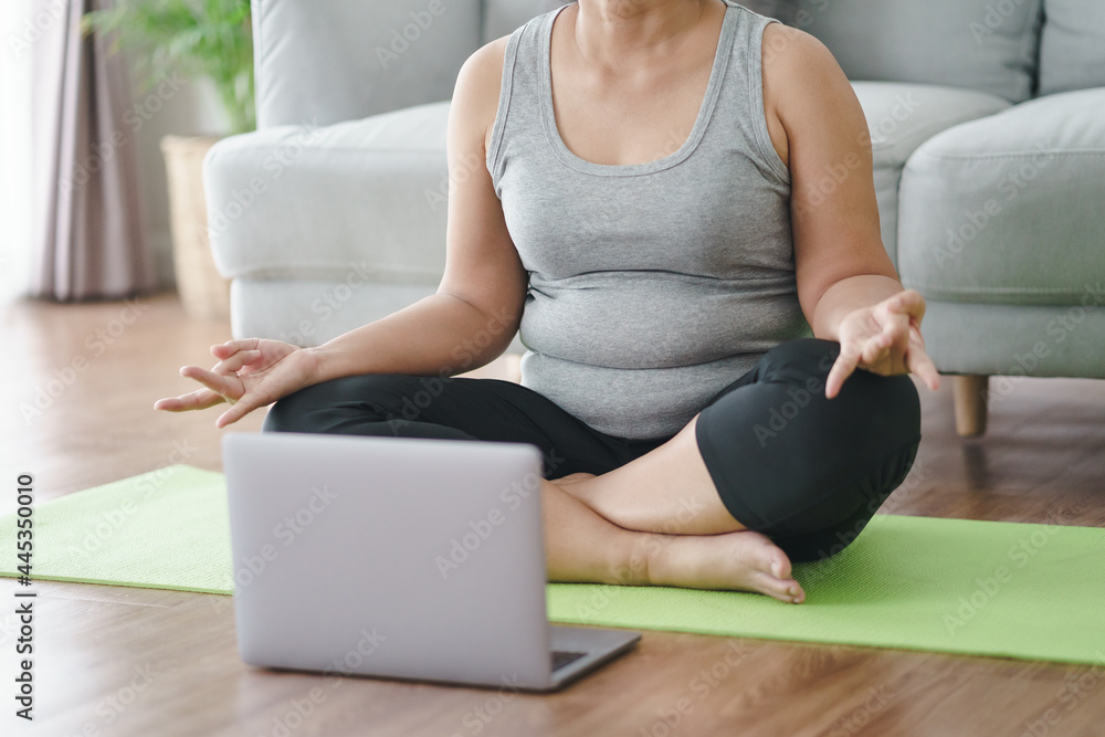 Fototapeta premium Mature Asian chubby fat woman sitting on the floor in living room practice online yoga lesson with the computer. female having meditate training class on the laptop..