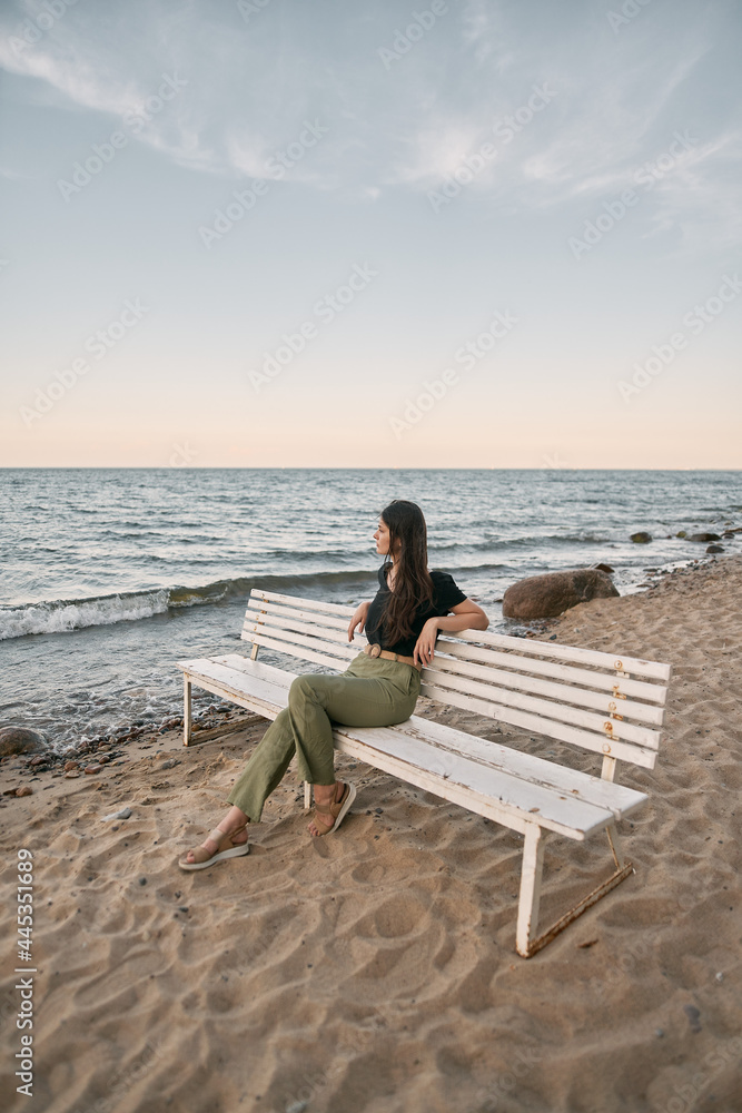 Beautiful brunette woman looking far into the sea horizon. Girls sits on white bench on the beach. Concept of work-life balance and wellbeing.