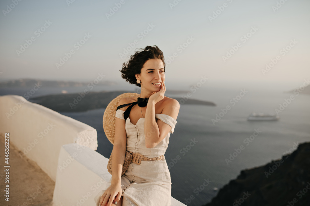 Brunette short-haired woman in elegant beige dress and boater enjoys beautiful sea view. Curly lady in good mood sits on white fence. Brunette short-haired woman in elegant beige dress and boater enjoys beautiful sea view. Curly lady in good mood sits on white fence.
