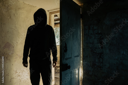 A horror concept. Of a hooded figure with no face standing in the doorway of a decaying room in an abandoned ruined house