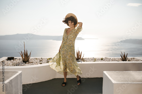Fotografie Joyful brunette woman in floral dress and hat smiles on ocean background
