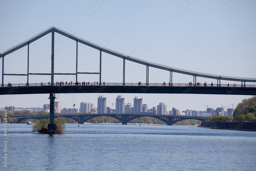 Fototapeta premium Parkovy Pedestrian Bridge about Dnieper river with ships passing beneath, Kiev, Ukraine. Silhouettes of people walking on the bridge. Bridge bunjee jumping. Extreme sport. River navigation.