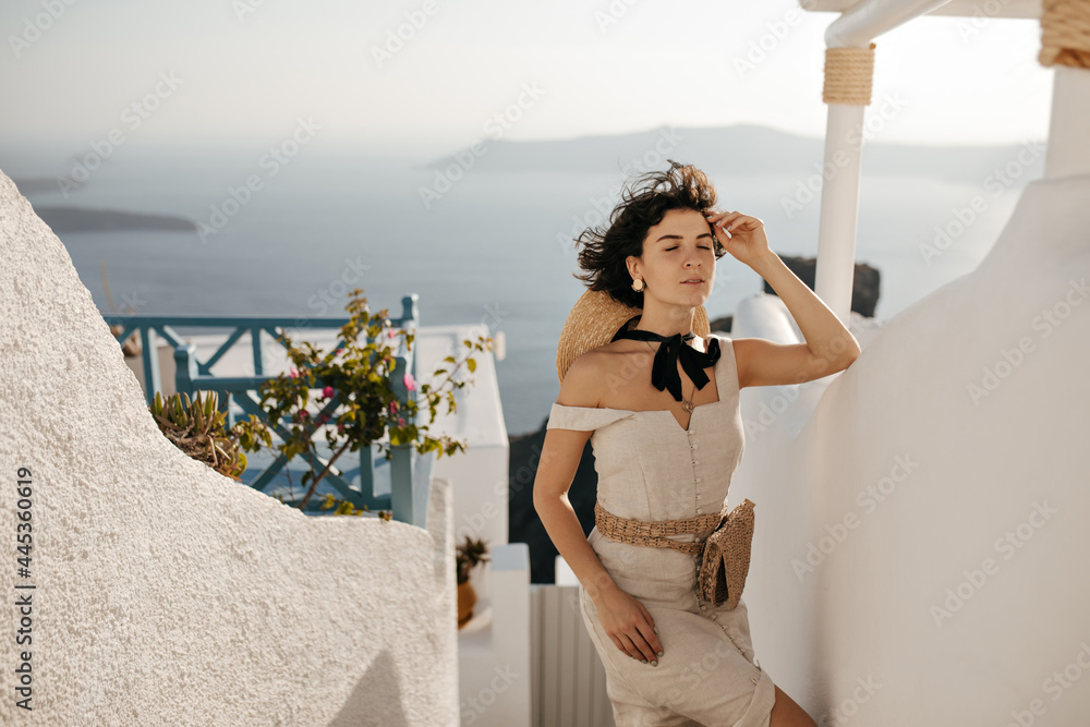 Pretty young brunette woman in beige summer dress poses in beautiful place with sea view. Curly lady in stylish outfit leans on white wall outside. - obrazy, fototapety, plakaty 