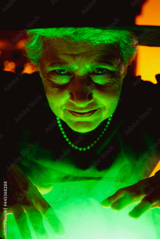 © Alberto - Smiling old woman in classic witch costume preparing green magic potions and looking at camera. senior person celebrating halloween