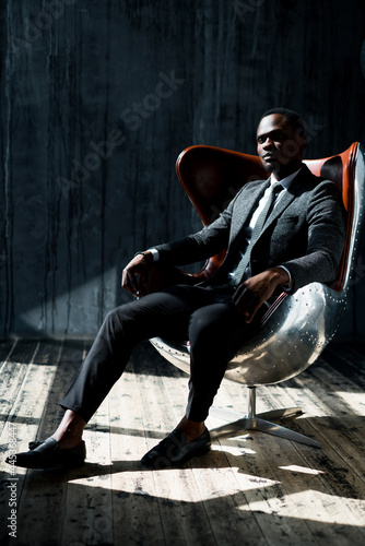 Stylish attractive african american businessman in a suit sits on a chair in the office and looks to the side