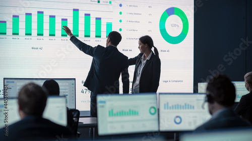 Two Traders Having a Meeting in a Modern Monitoring Office with Analytics Feed on a Big Digital Screen. Monitoring Room with Brokers and Finance Specialists Sit in Front of Computers. Colleagues Talk. photo