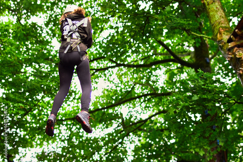 Young woman in safety equipment harness climbing and jumping from tree leap of faith on rope space for text