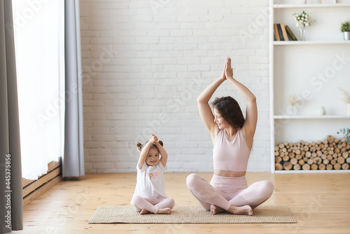 woman looking at her small kids girl sitting on the carpet floor in lotus pose namaste hands up