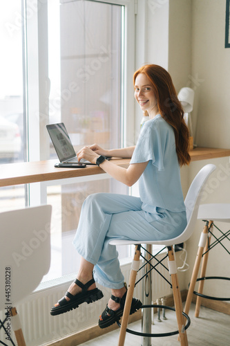 Cheerful young woman wearing casual clothing typing on laptop computer sitting at table by window in cafe, looking at camera. Pretty redhead Caucasian lady remote working or studying. © dikushin