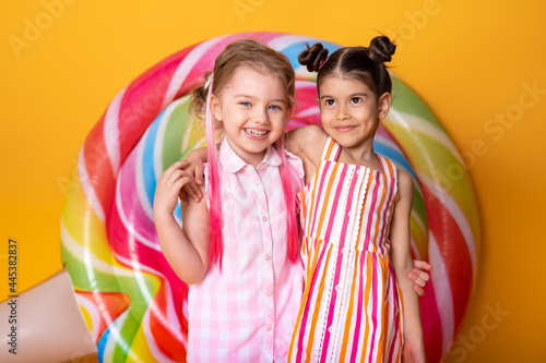 two happy little girls in colorful dress laughing hugging having fun on yellow background with lollipop