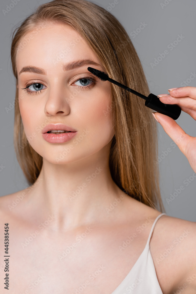 young woman holding brush and applying mascara isolated on grey