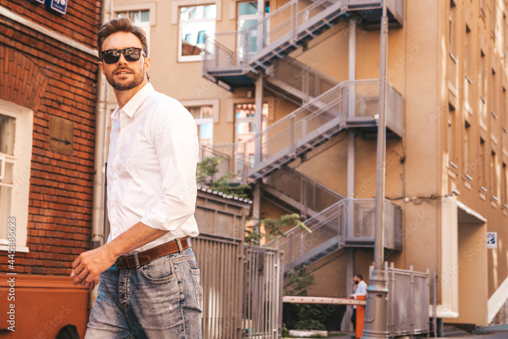 Portrait of handsome smiling stylish hipster lambersexual model.Modern man dressed in white shirt. Fashion male posing in the street background near skyscrapers in sunglasses. Crossed arms