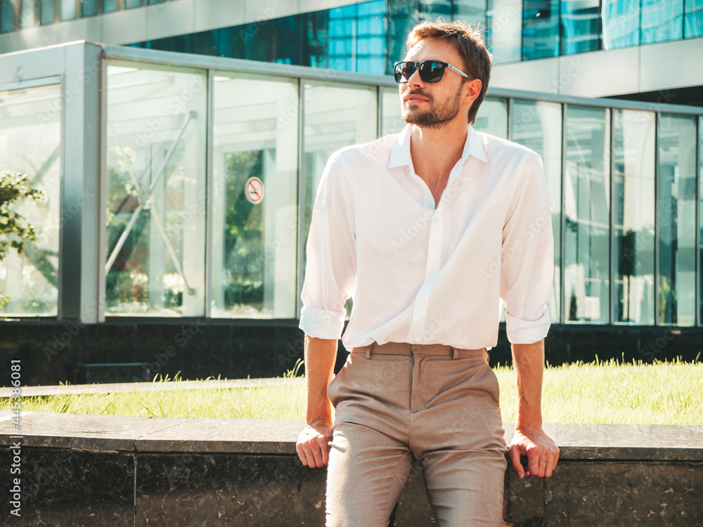 Portrait of handsome smiling stylish hipster lambersexual model.Modern man dressed in white shirt. Fashion male posing in the street background near skyscrapers in sunglasses