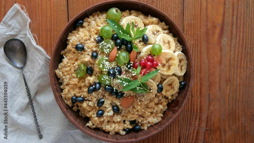 Wholegrain Oatmeal porridge with fresh berries, fruits nuts mint leaves sesame seeds in female hand. Healthy food oat meal breakfast top view. Whole grain cereal gooseberry currant banana wooden table Wholegrain Oatmeal porridge with fresh berries, fruits nuts mint leaves sesame seeds in female hand. Healthy food oat meal breakfast top view. Whole grain cereal gooseberry currant banana wooden table