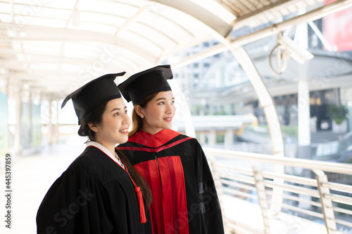 Two Asian women wore graduate gowns are looking forward and smiling widely.