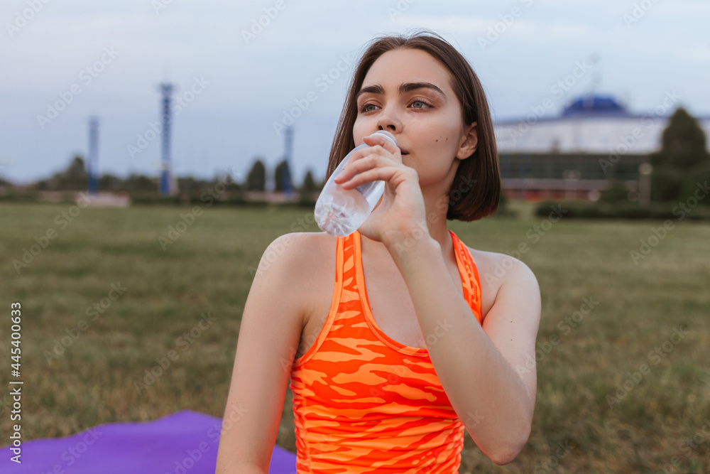 Fit thirsty sportswoman in bright activewear drinking fresh water from bottle during training in field in city Fit thirsty sportswoman in bright activewear drinking fresh water from bottle during training in field in city