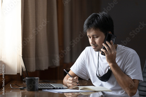 Young Asian man talking on the phone and make notes sit at work desk, concept work from home