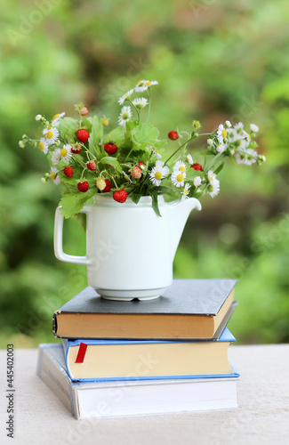 A strawberry bush in a teapot and a bouquet of daisies and a seashell on a stack of books on a table in the garden. Summer Leisure and Relaxation. Vertical photo background