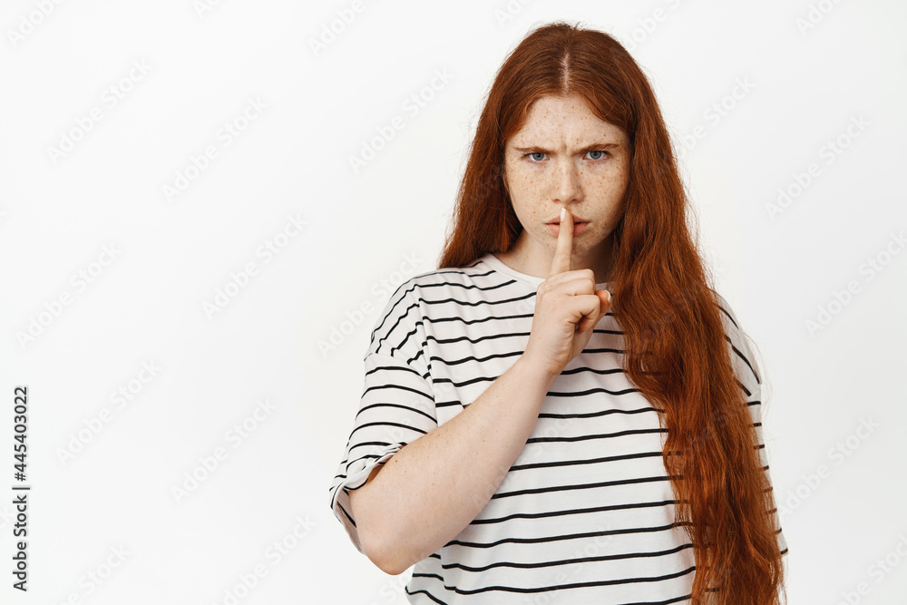 Angry redhead teen girl hush, frowning upset and shushing, tell to be quiet, scolding loud behaviour, warning, showing taboo sign, standing against white background
