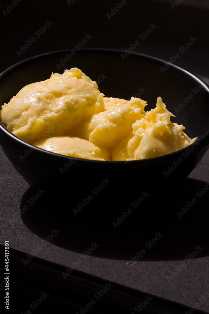 Rich creamy textured butter closeup view from the front. The golden yellow butter served in black ceramic bowl, placed on black background, lit up by harsh sunlight shines,glows and casts deep shadows Rich creamy textured butter closeup view from the front. The golden yellow butter served in black ceramic bowl, placed on black background, lit up by harsh sunlight shines,glows and casts deep shadows