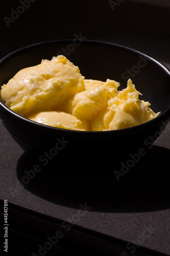 Rich creamy textured butter closeup view from the front. The golden yellow butter served in black ceramic bowl, placed on black background, lit up by harsh sunlight shines,glows and casts deep shadows