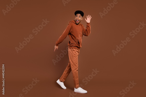 Full length profile photo of funky short hairdo young guy wave empty space dress sportswear trousers sneakers isolated on brown color background