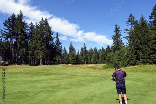 A young male golfer playing alone, standing in the middle of the fairway measuring his distance, surrounded by forest, on a beautiful sunny day in Campbell River, British Columbia, Canada.