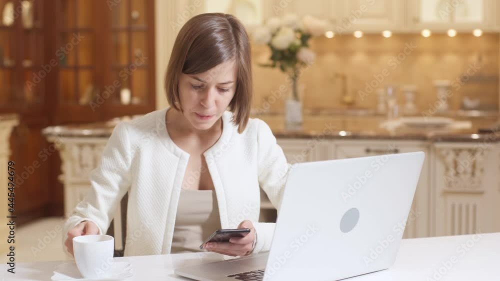 Business Woman In White Clothes Sitting At Desk With Laptop Computer Drinking Coffee And Uses Mobile Phone at Home Office Workplace at Modern Kitchen. Girl Female Freelancer Chatting On Phone. Business Woman In White Clothes Sitting At Desk With Laptop Computer Drinking Coffee And Uses Mobile Phone at Home Office Workplace at Modern Kitchen. Girl Female Freelancer Chatting On Phone.