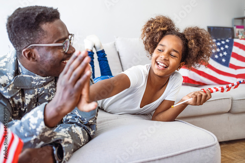 Portrait of happy american family father in military uniform and cute little girl daughter with flag of United States hugging and smiling at camera, male soldier dad reunited with family at home. 