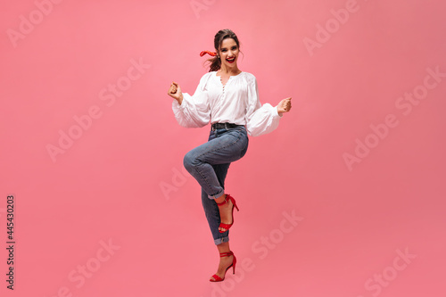 Joyful woman in jeans, white blouse dancing on pink background. Modern girl with red lipstick and in stylish heels rejoices on isolated backdrop..
