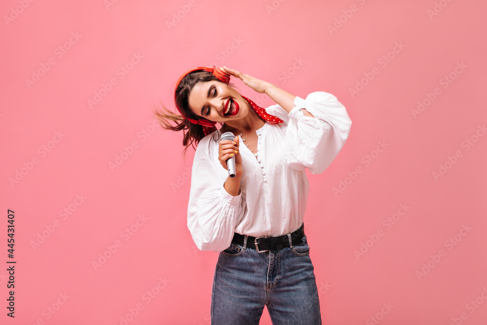 Dark haired lady in white stylish blouse and jeans listens to song in headphones and sings in microphone on isolated background.. Dark haired lady in white stylish blouse and jeans listens to song in headphones and sings in microphone on isolated background..