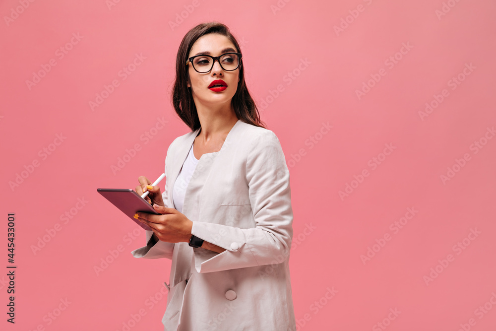 Woman in eyeglasses makes notes in tablet. Beautiful brunette with big red lips in light suit posing on isolated backdrop.. Woman in eyeglasses makes notes in tablet. Beautiful brunette with big red lips in light suit posing on isolated backdrop..