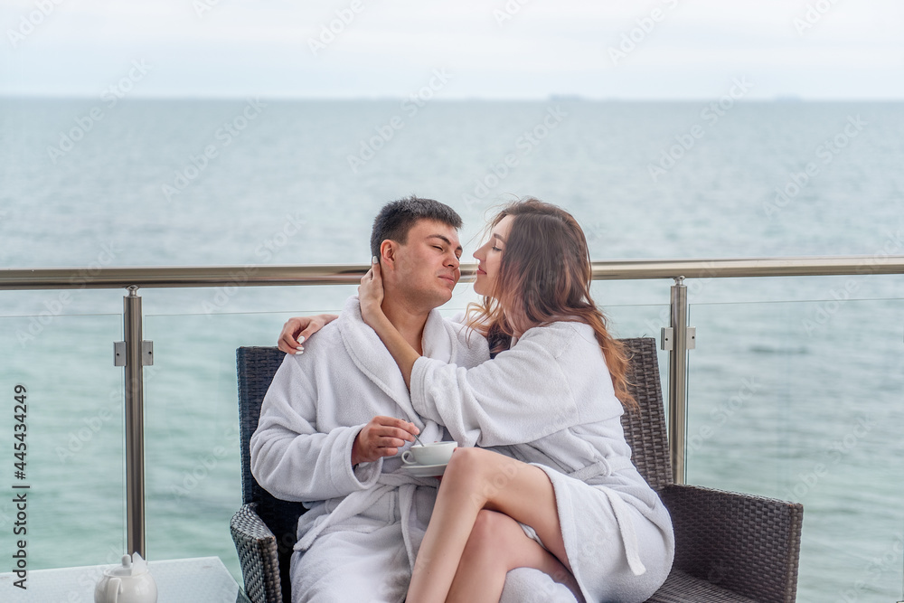 A couple in white bathrobes are sitting on an open terrace by the sea, drinking tea with lemon from cups. Lovers travel on vacation. Young man and woman hug each other. Newlyweds on their honeymoon