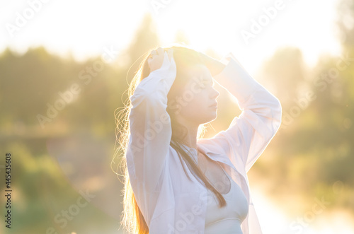 A beautiful red-haired girl in a white shirt meets the summer dawn in nature. The concept of happiness and spiritual freedom.